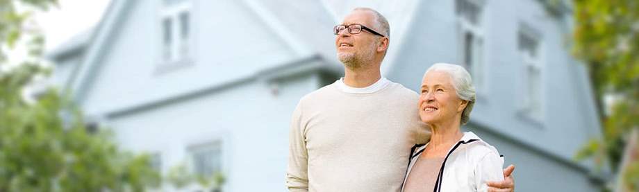 A senior couple stands in front of a blue house asking should I pay off my mortgage before I retire